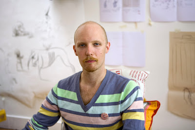 A picture of Alexis Deacon sitting on a chair in a room with papers stuck on the wall. he is wearing a multicoloured striped jumper and is sporting a close cropped shave and a a goattee.